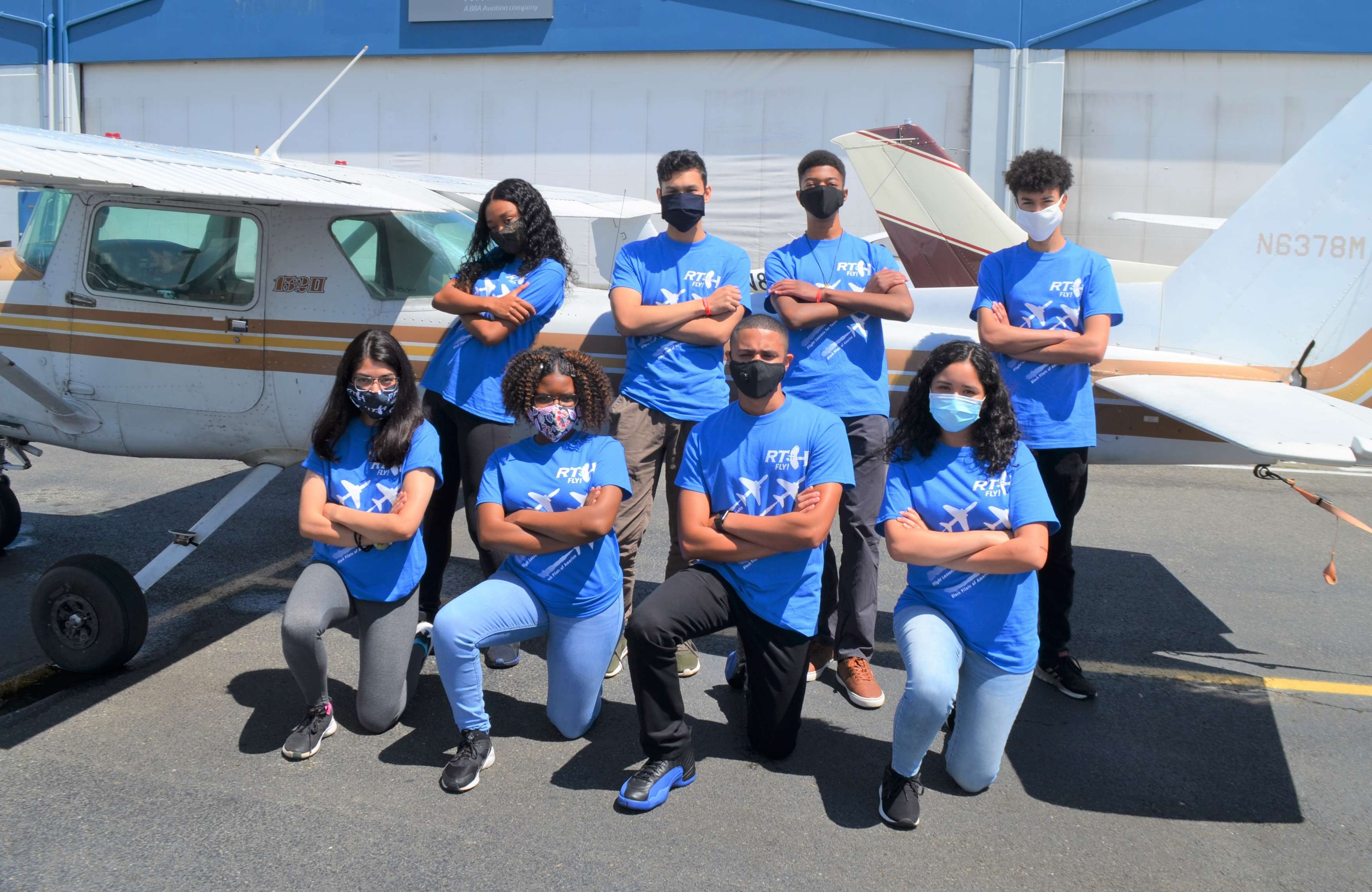 Eight youth wearing blue shirts pose in front of an airplane with their arms crossed