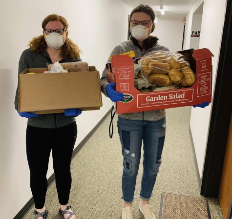 Two people wearing face masks and blue gloves stand in a hallway holding boxes of food. 