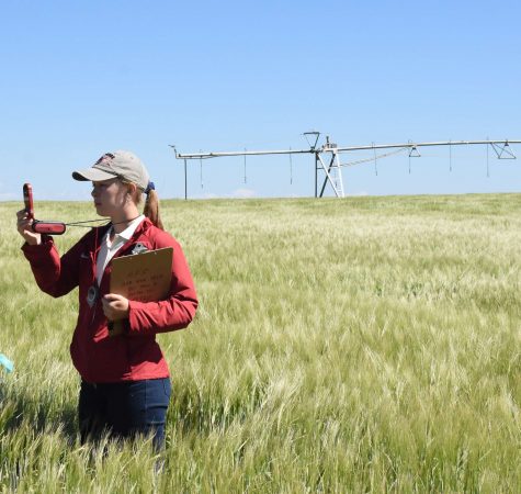 A woman wearing a red jacket and tan hat holds a clipboard and collects data while standing in a field. 