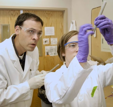 A man with short dark hair wearing a white lab coat and protective goggles and a woman with straight brown hair wearing a white lab coat, protective goggles, and purple gloves observe something in a lab. 