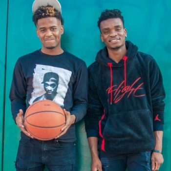 Two young Black men pose in front of a blue wall, one of them holding a basketball
