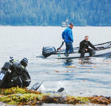 A man sits on the shore of Sitka Sound in a scuba suit while two men ride by in a raft