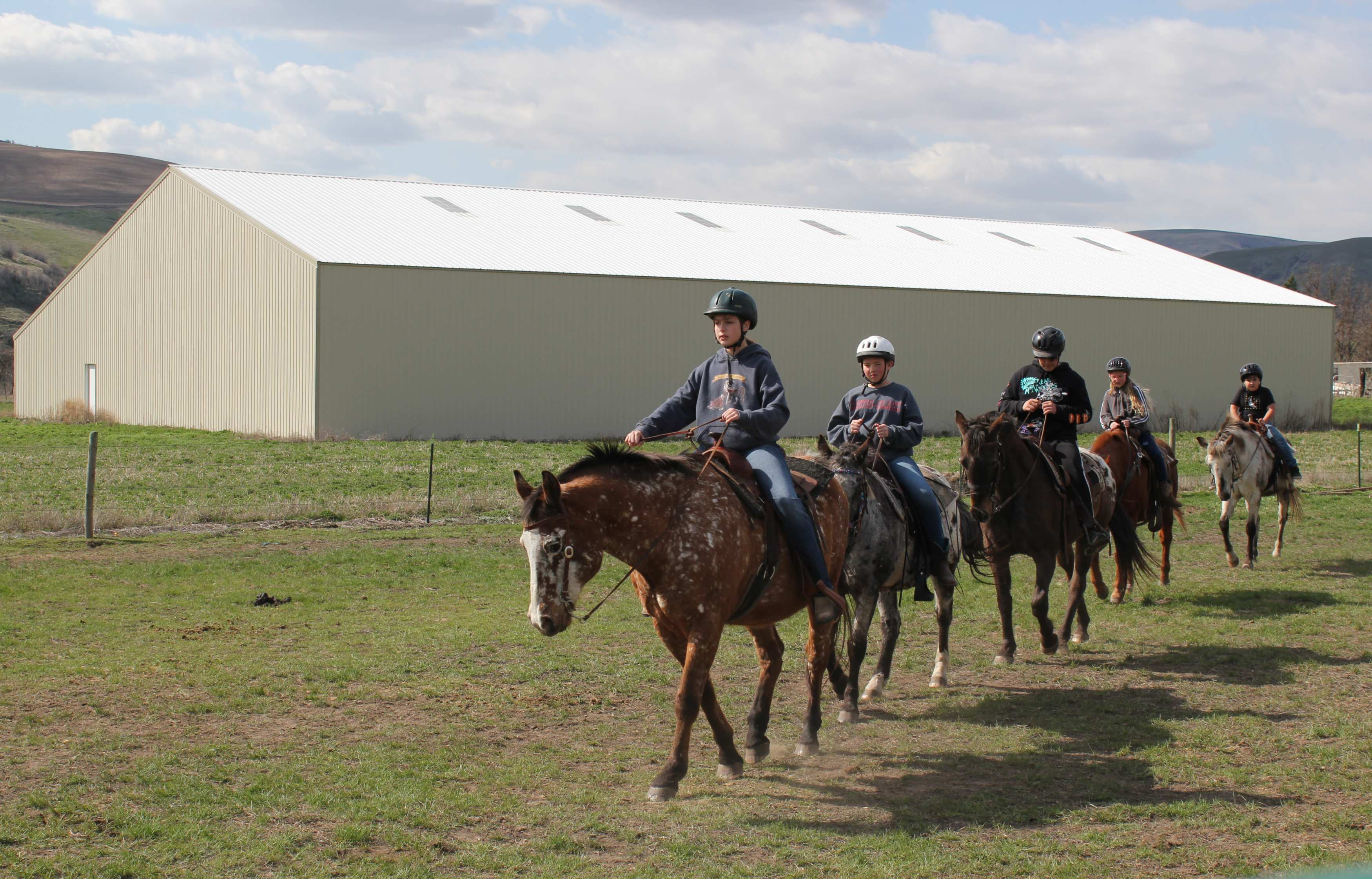 Five Appaloosa horses with riders on them by a white riding facility.