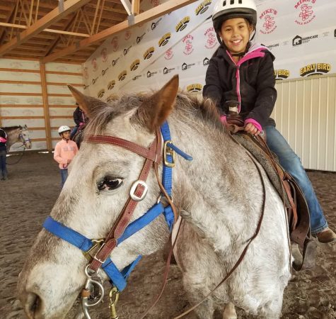 A young girl wearing a white helmet sits on a white Appaloosa horse and smiles for the camera inside a horse riding arena.