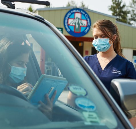 A woman with straight brown hair in a ponytail wearing a blue mask and a Girdwood Health Clinic shirt waits for someone to fill out a tablet in their car.