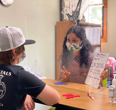 A woman with dark curly hair wearing a mask talks to a young boy wearing a hat through plexiglass