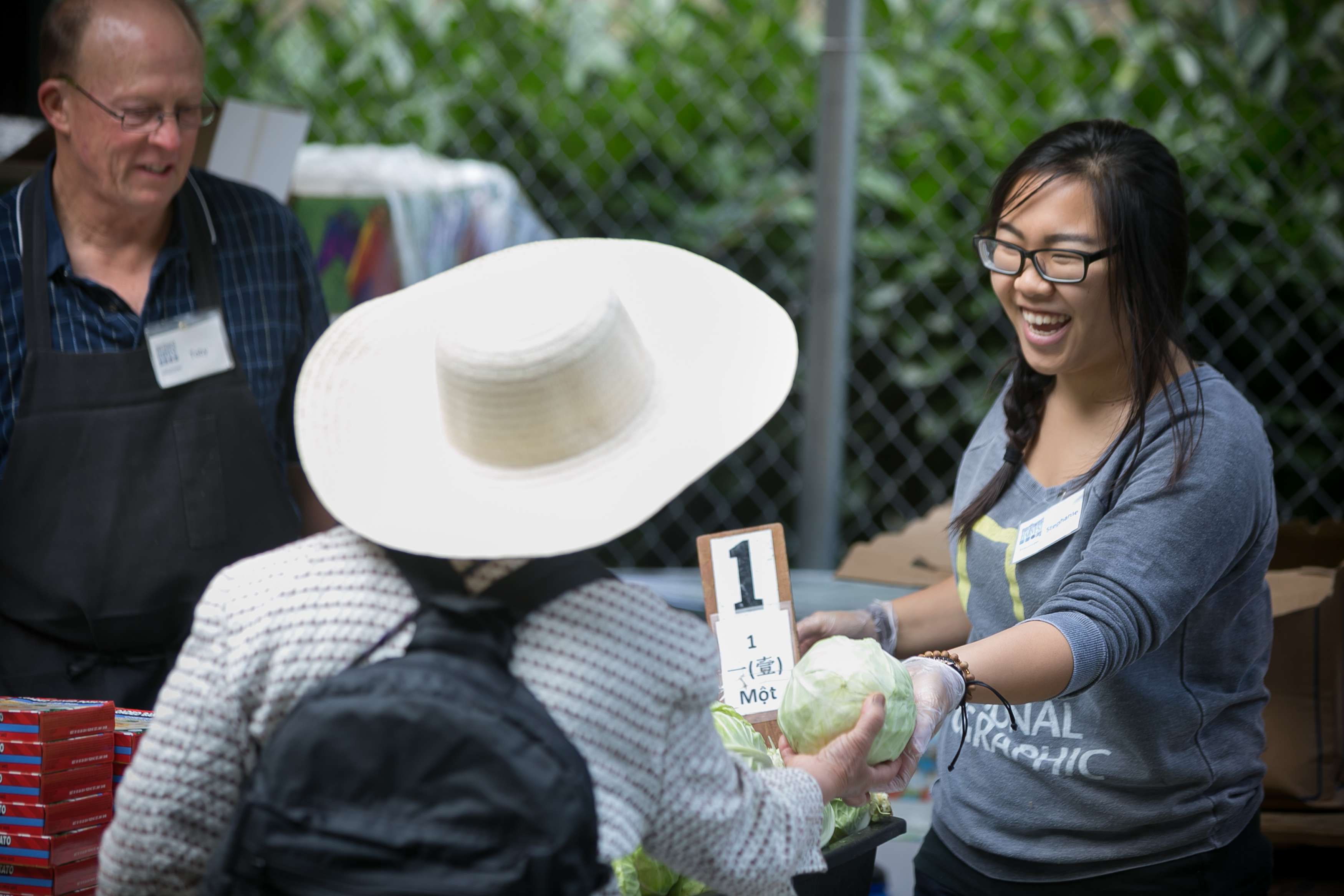 A woman with dark straight hair wearing glasses hands a vegetable to a woman wearing a straw hat at a produce line.