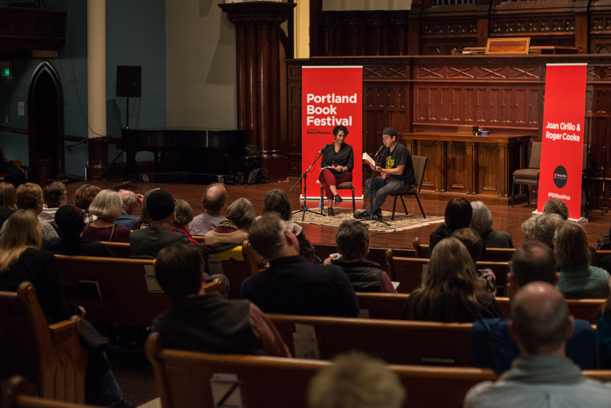 Two people from Literary Arts sit on stage in front of red banners that read "Portland Book Festival" in front of a large audience