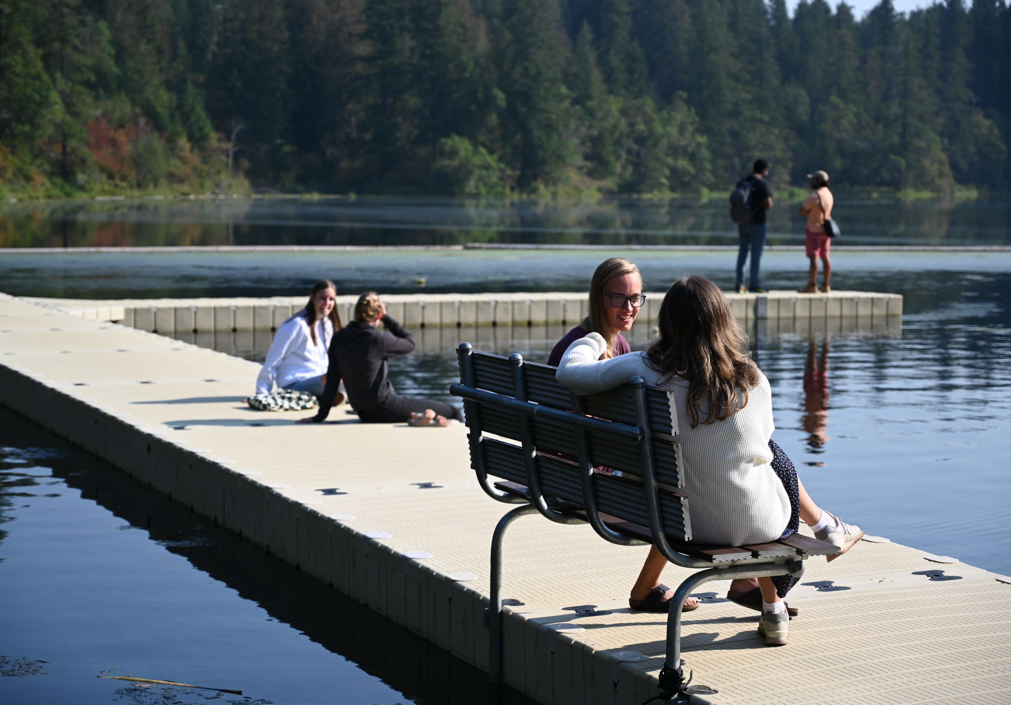 Two young women talk to each other on a bench near a lake, with two other sets of people on the dock behind them. Pine trees are in the distance. This is from the Vision & Call 2022 Retreat.