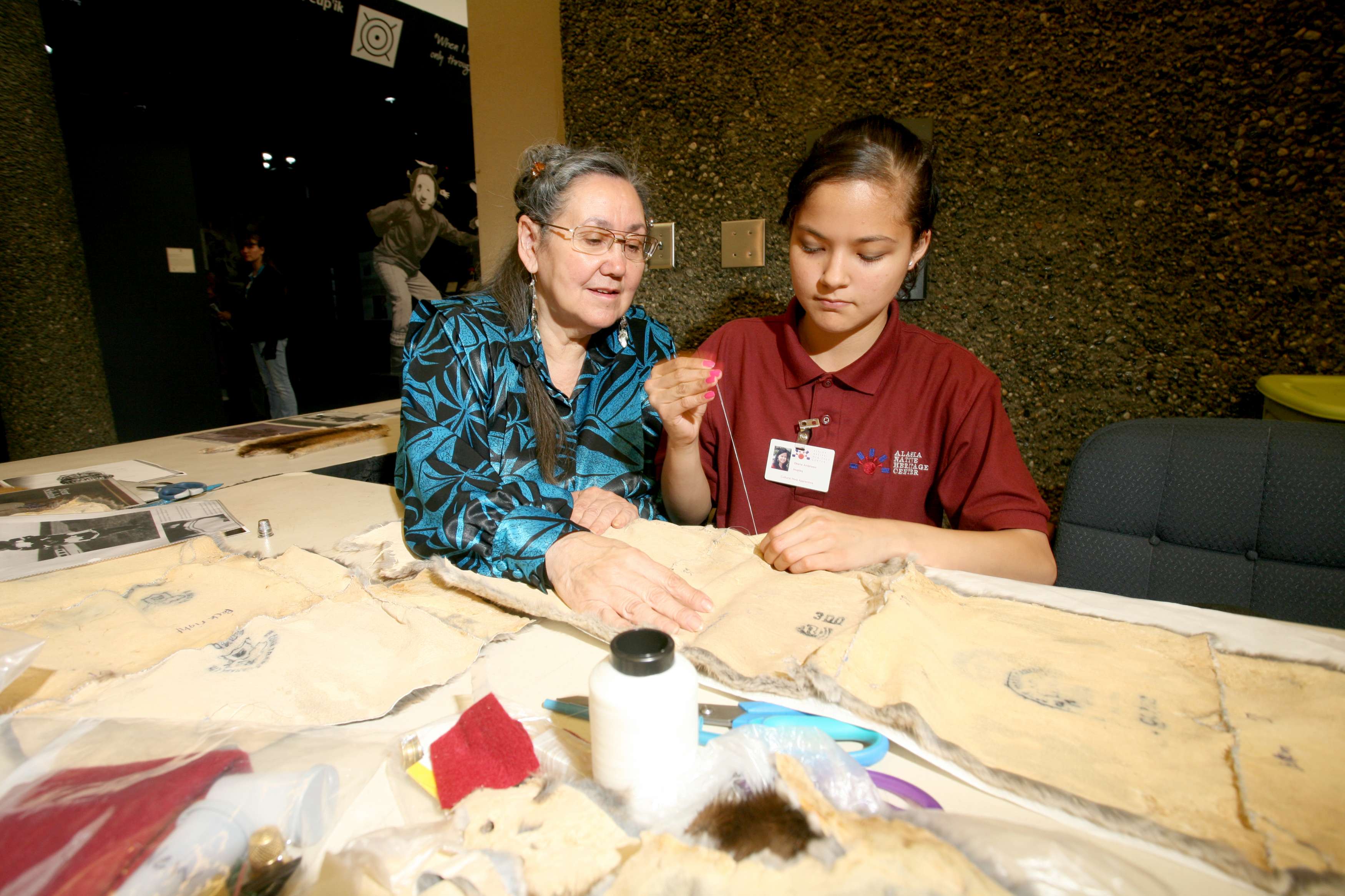 A woman helps a young girl make traditional Native art using a thread and needle.
