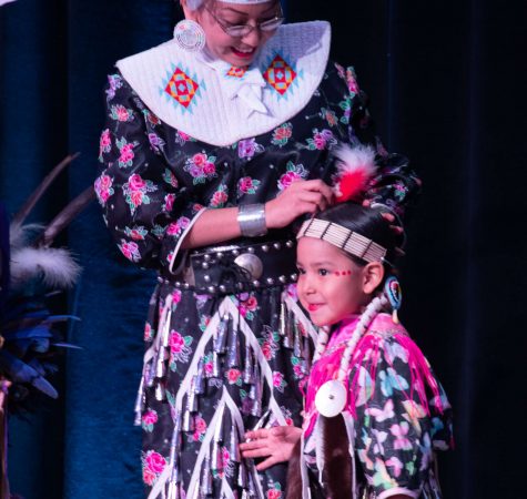A young girl wearing a headdress and colorful clothing smiles near a women fixing her hair. 