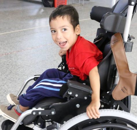 A child wearing a red shirt and blue pants smiles at the camera while riding in a wheelchair.