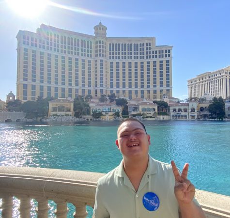 A young man wearing a blue Make-A-Wish pin smiles in front of the Bellagio Fountains. 