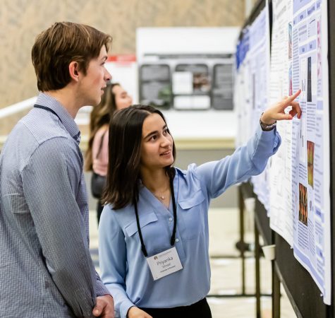 A woman with dark hair wearing a blue shirt explains something to a man with brown hair while pointing to her poster presentation at the 2022 MCSR Conference.