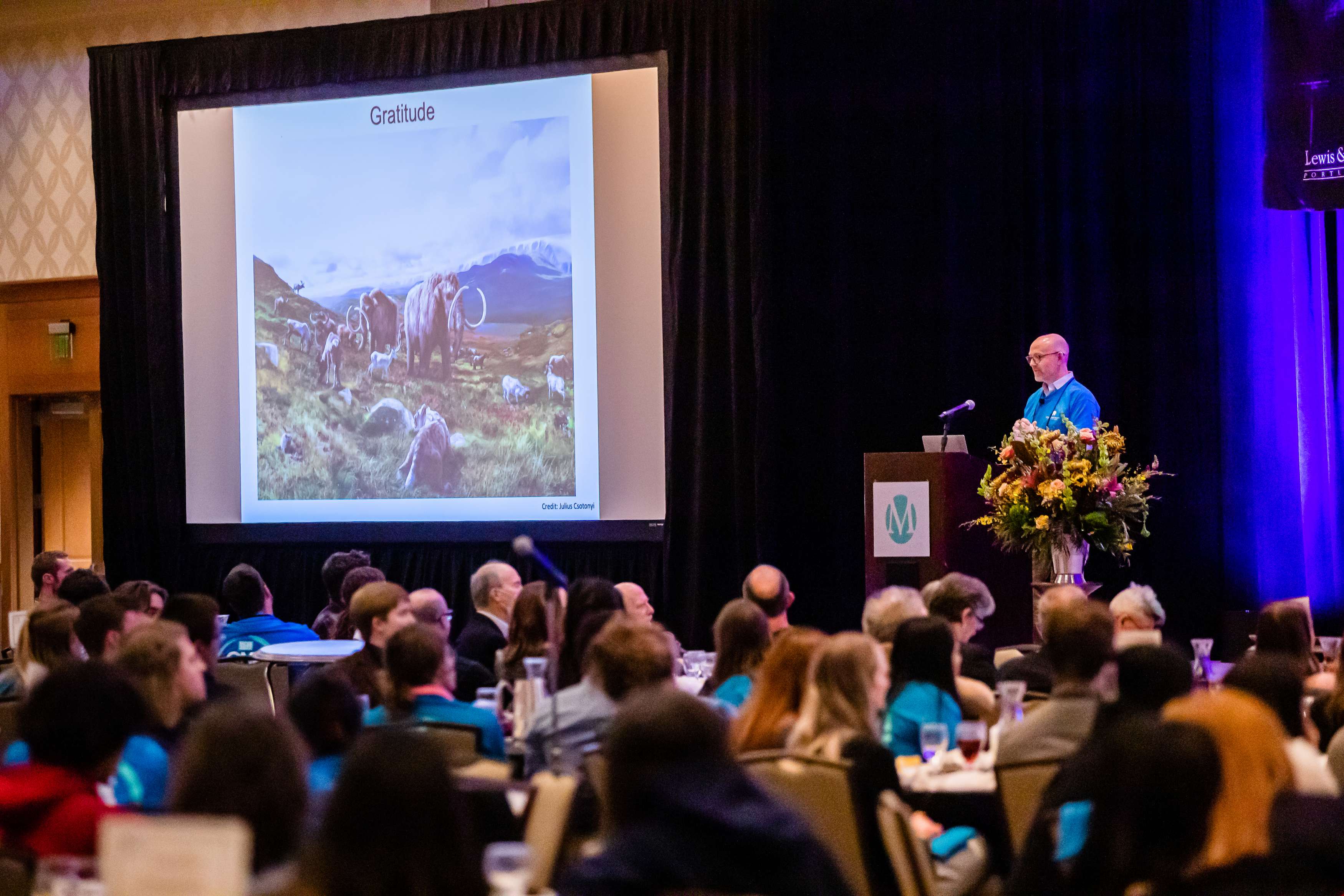 A man wearing a blue shirt presents a lecture at the 2022 MCSR Conference, with a large screen showing a picture of a woolly mammoth.