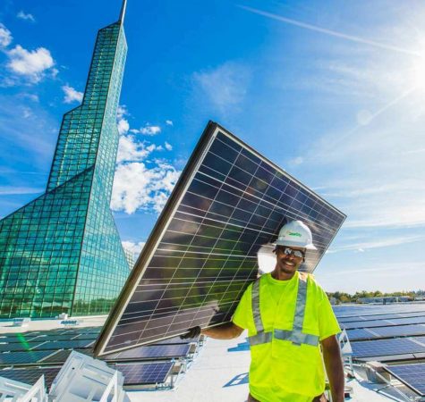 A man wearing a reflective shirt and construction hard hat carries a solar panel on top of a roof.