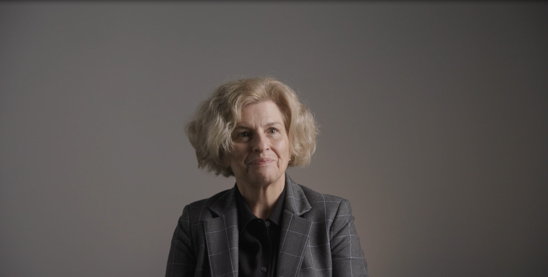 A woman with short blond hair wearing a gray blazer looks at the camera in front of a gray backdrop.