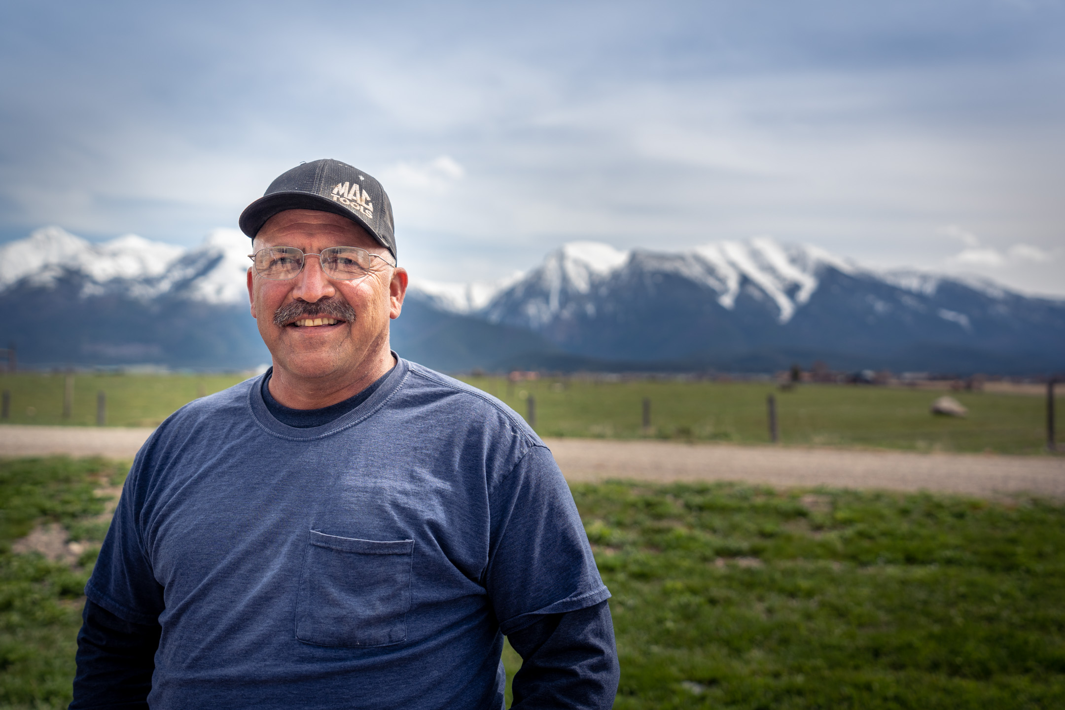 A man wearing a blue shirt and hat smiles for the camera, with mountains and green grass in the background.