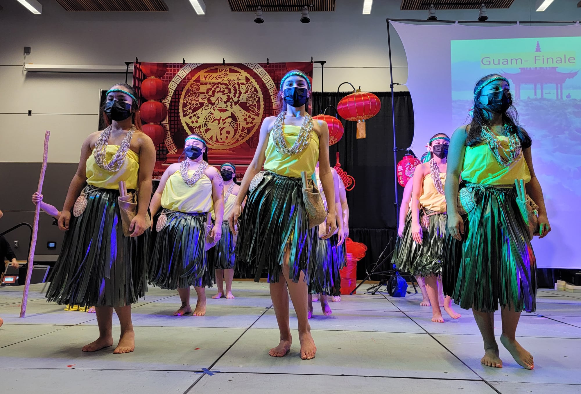 A group of individuals wearing hula skirts, necklaces, and headbands dance inside a room.