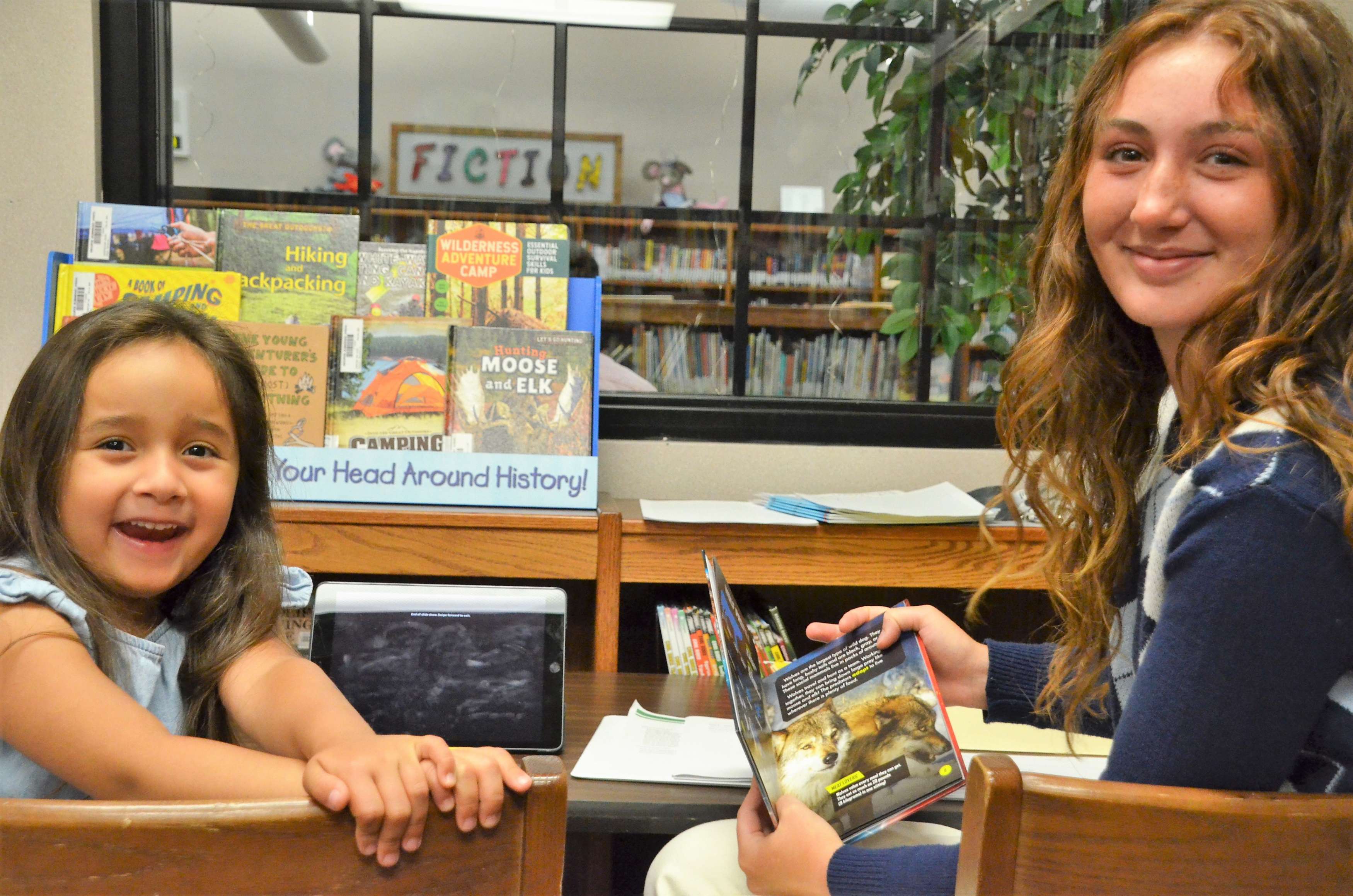 A young student and a volunteer smile at the camera while they read a picture book.