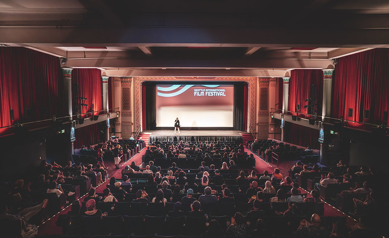 A full theater with red curtains and a screen that says "Seattle International Film Festival" with a person on stage speaking into a microphone