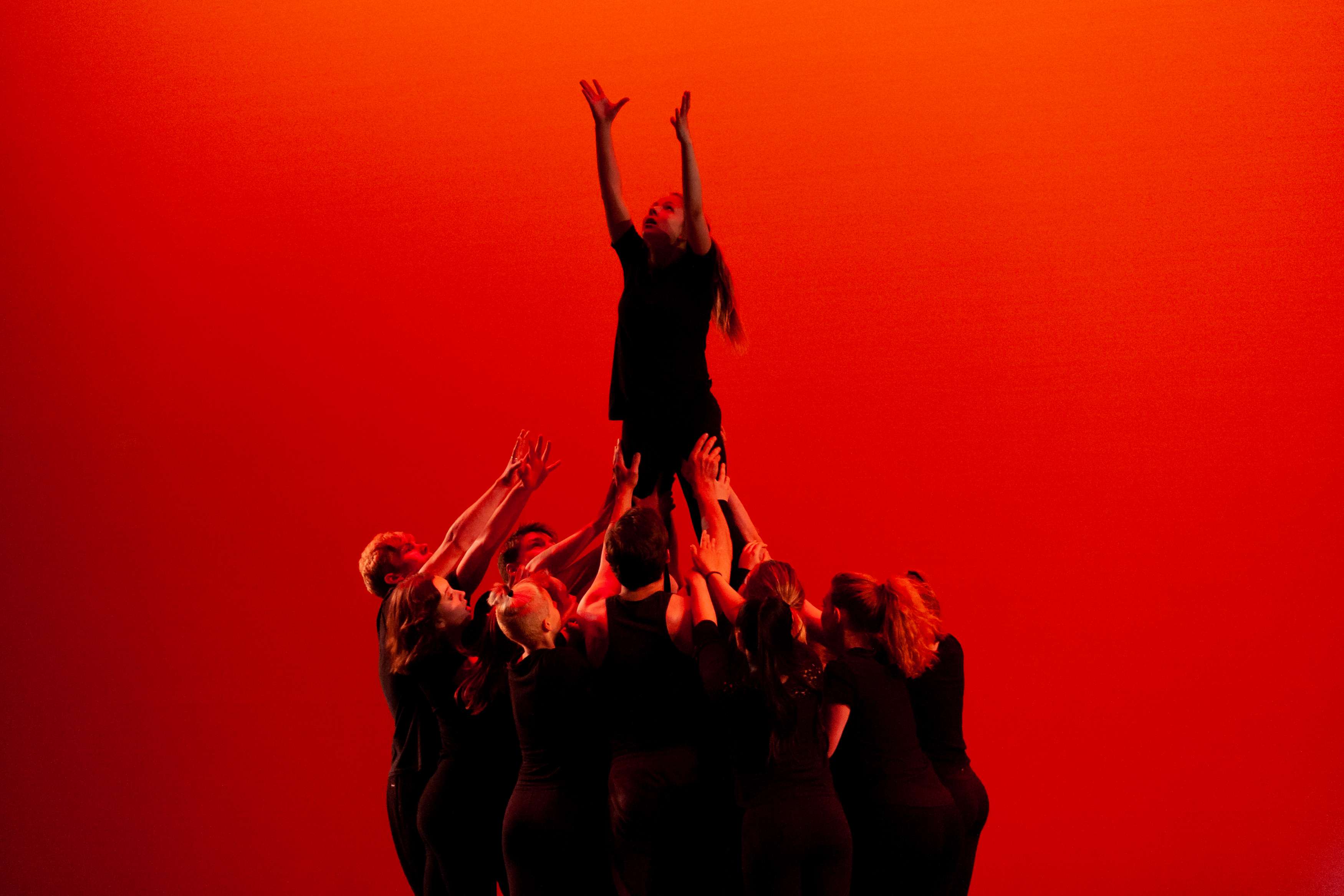 A group of dancers lifts another dancer with her arms raised toward the sky, against a red backdrop