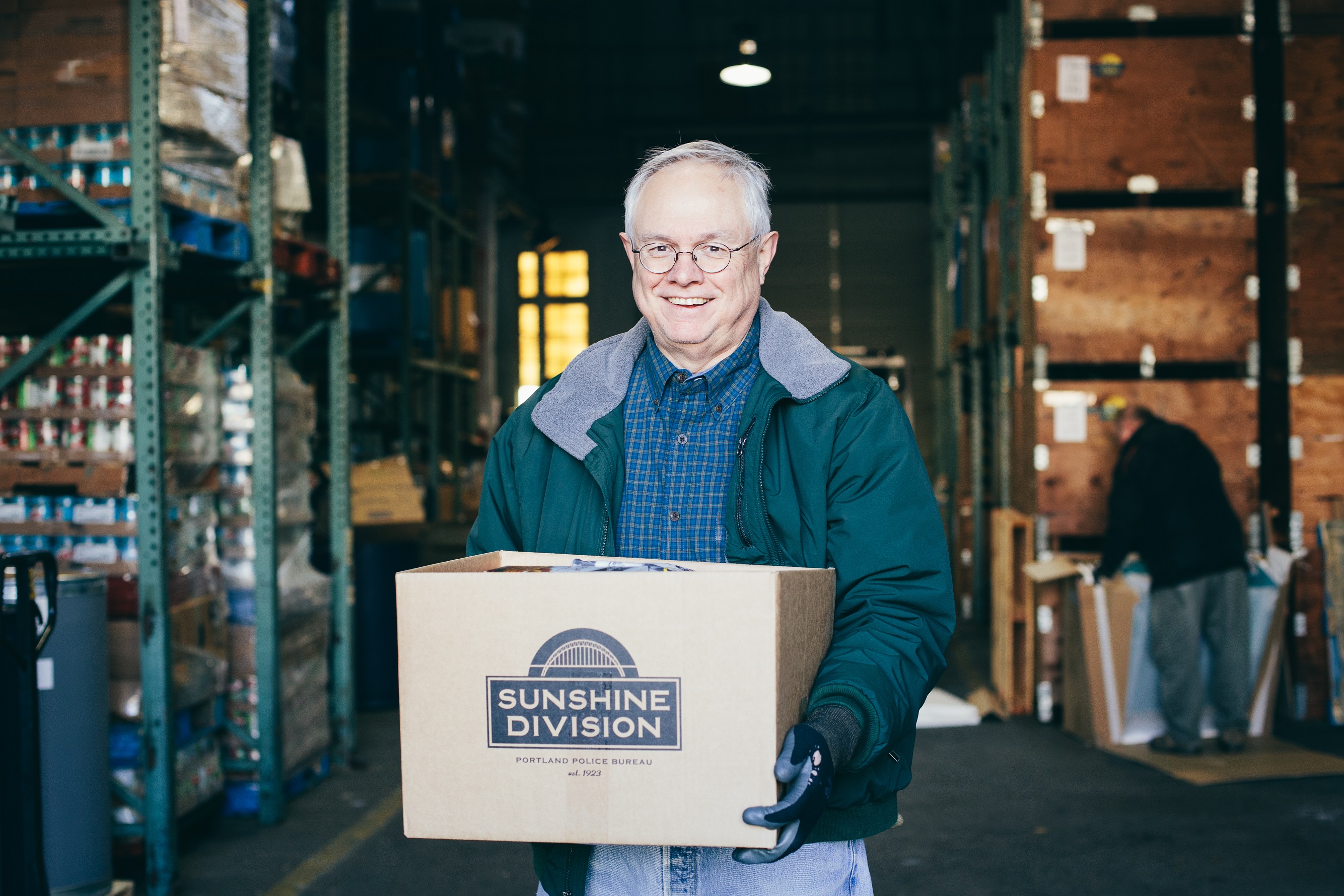 A man smiles and holds a box that says "Sunshine Division" inside a warehouse