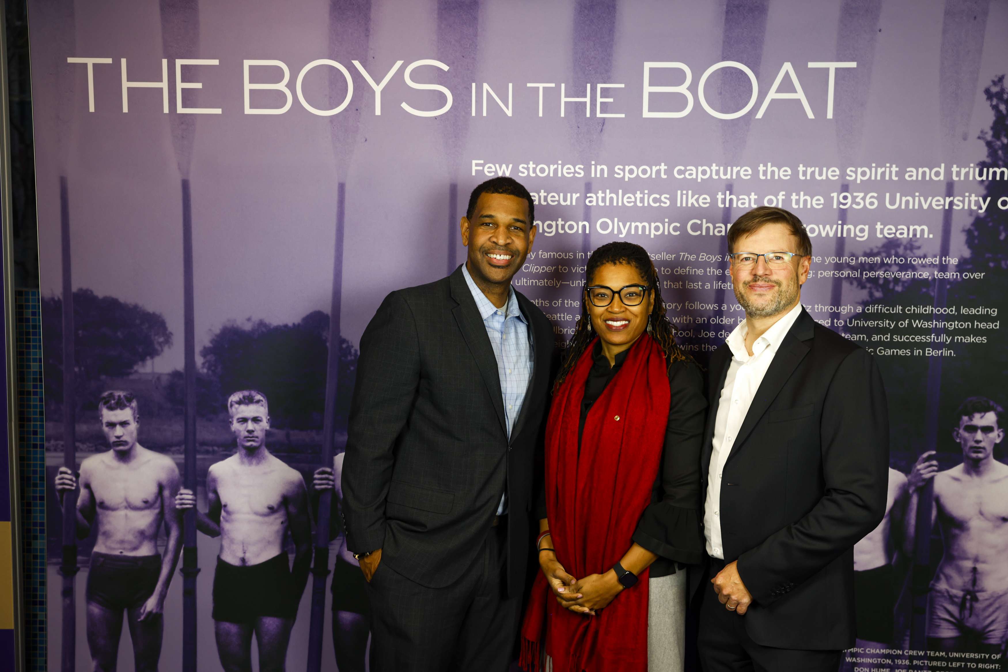 Three individuals pose in front of a sign that says "The Boys in the Boat" at the University of Washington