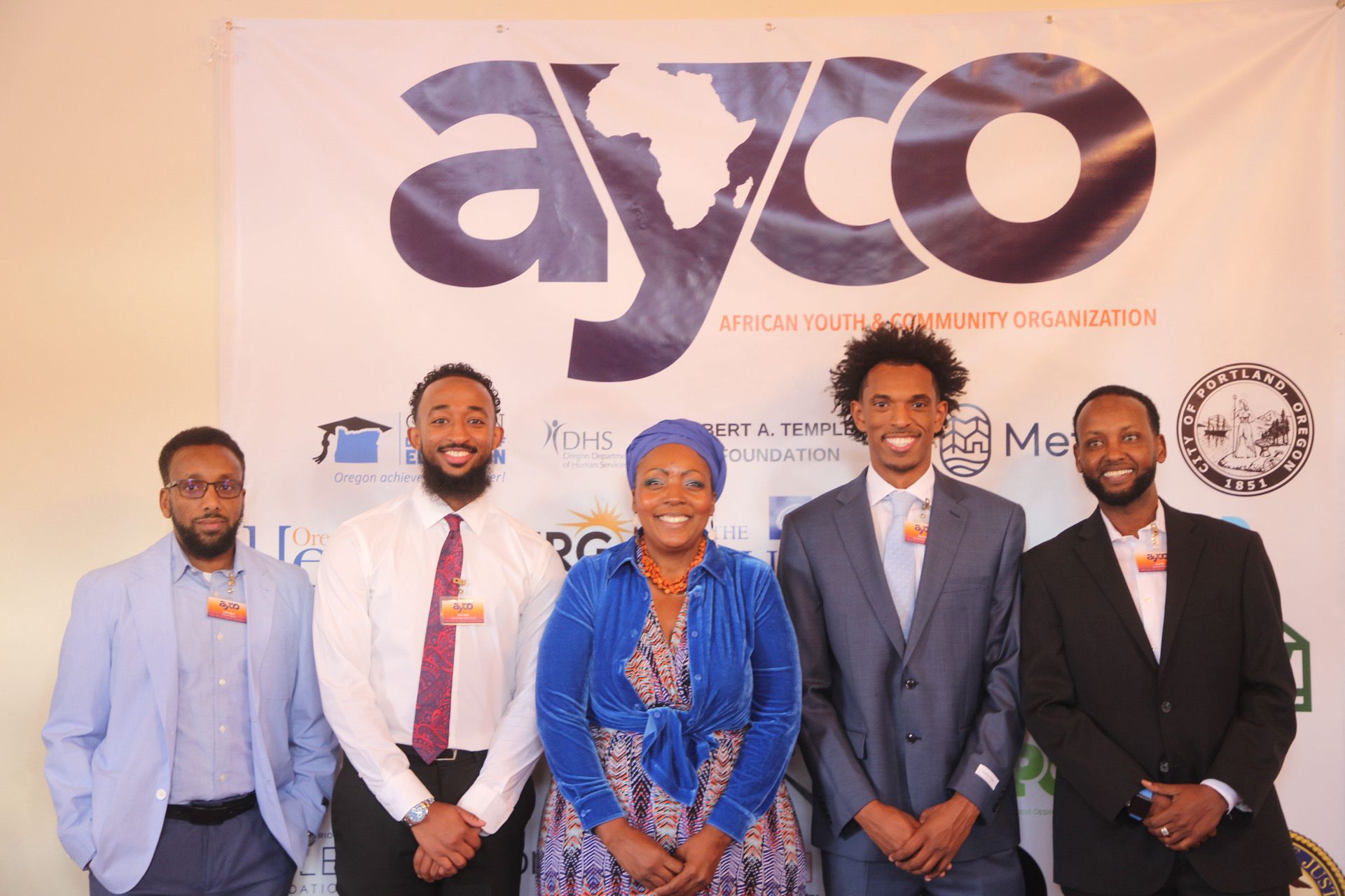 Four men and one woman dressed in business professional attire standing in front of an AYCO sign smiling 