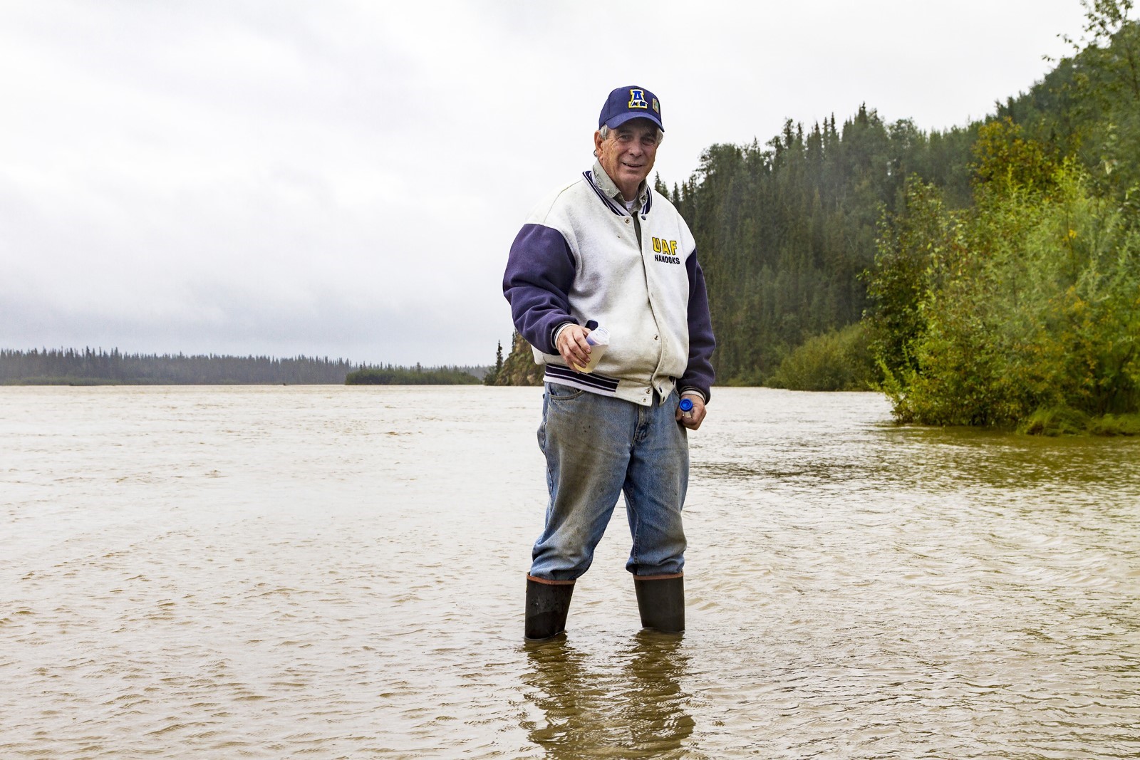 Man standing in a river, wearing waders and holding a fishing rod. They are wearing a jacket and hat with the logo 'Bass Pro Shops'. The background features a forest and overcast sky.