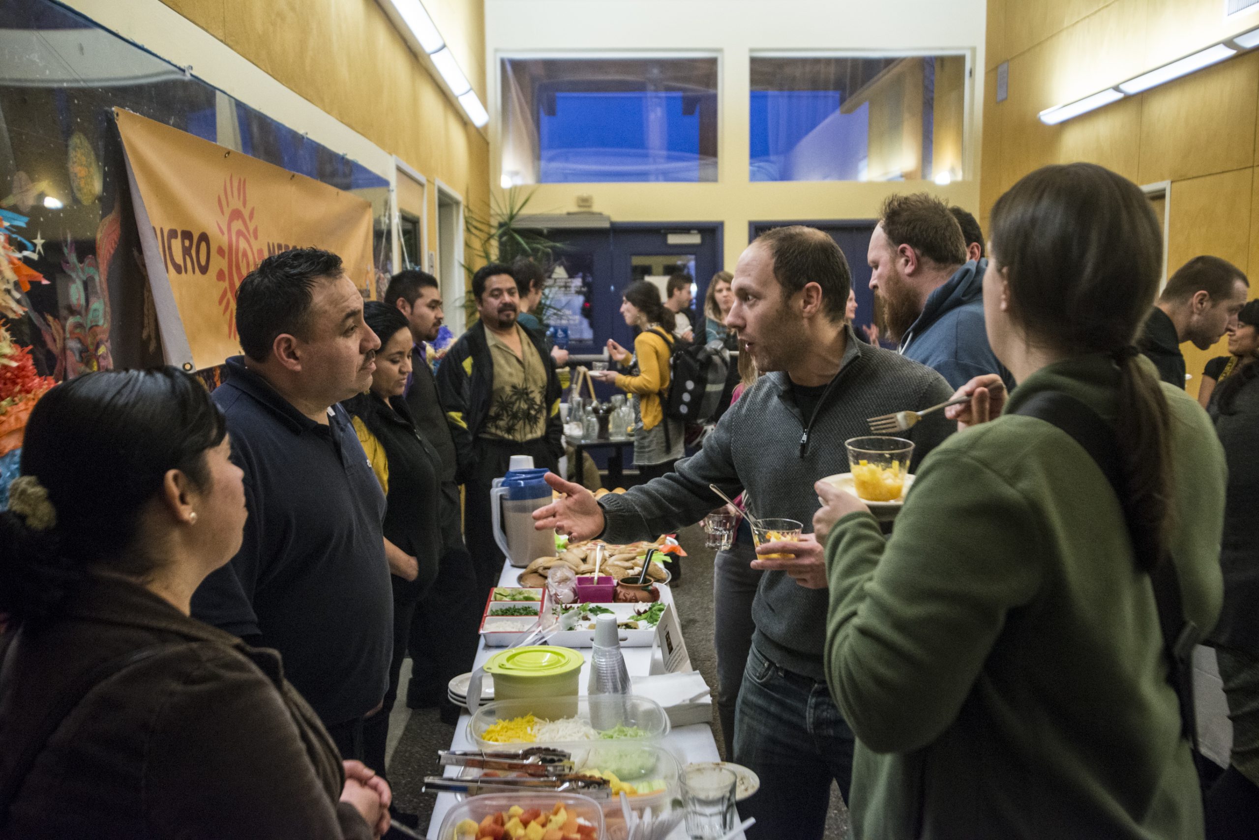 Long tables inside building with food. People serving up the food and on the other side of the table people dishing up