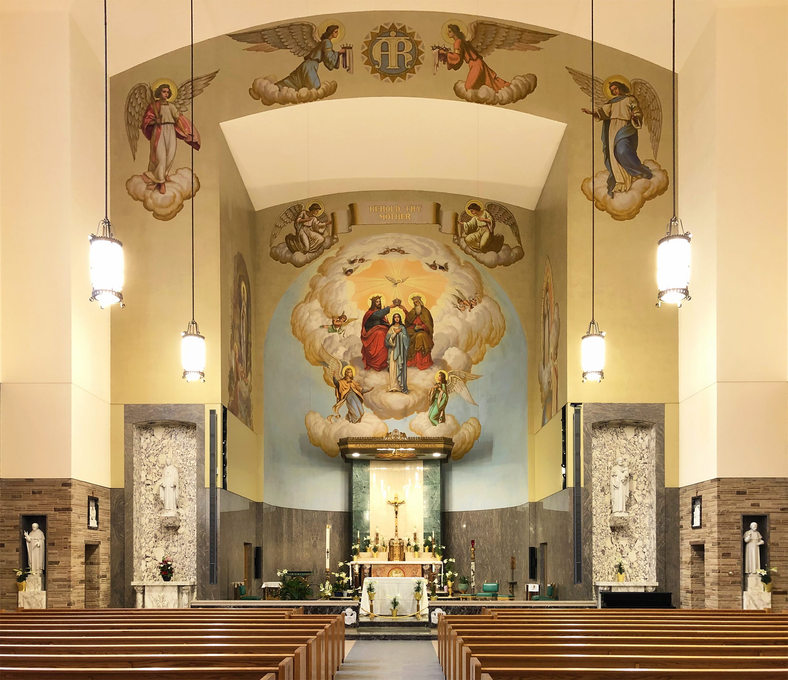 Interior of a church showing an altar with religious artwork above, including depictions of angels and holy figures. The church features rows of pews, hanging lights, and a serene ambiance.