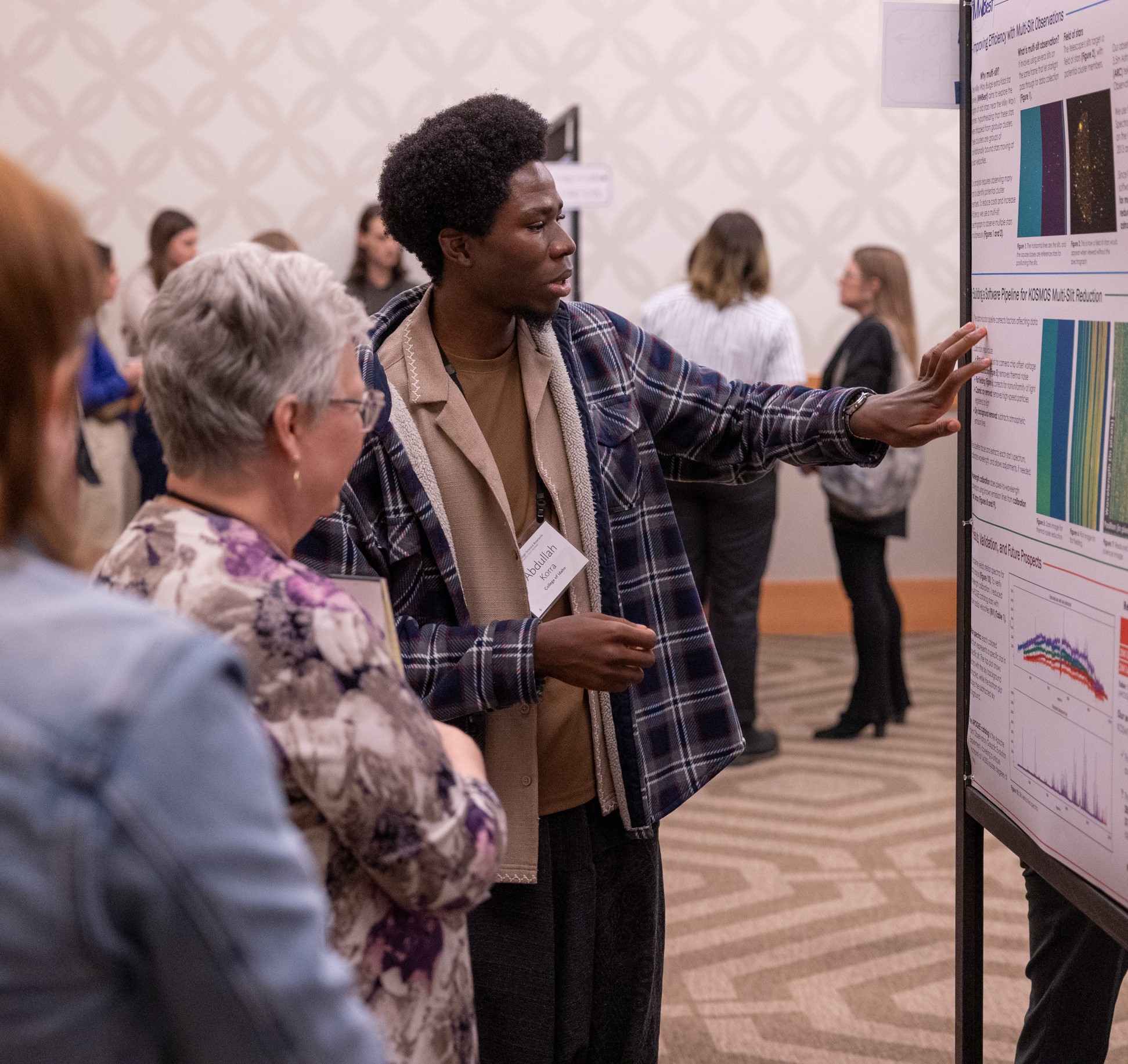 Student presenting his research and gesturing towards hist poster with two people intently listening