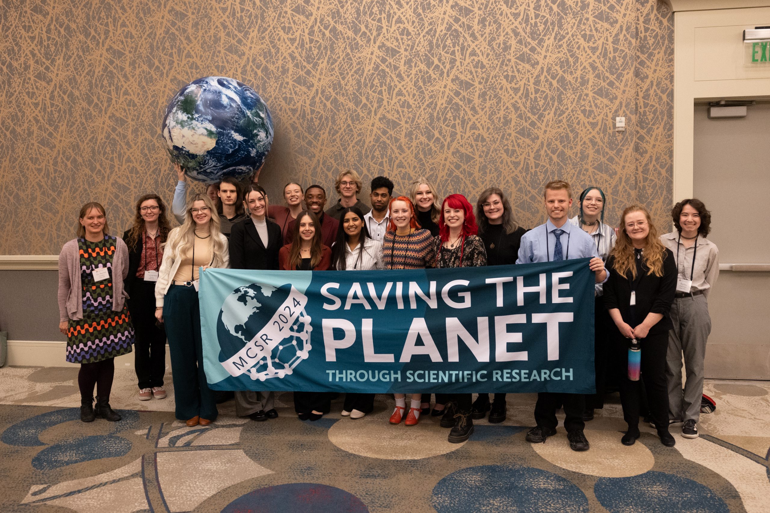 Students smiling and standing in a conference room. People in front holding up a large sign that says, "Saving the Planet Through Scientific Research"