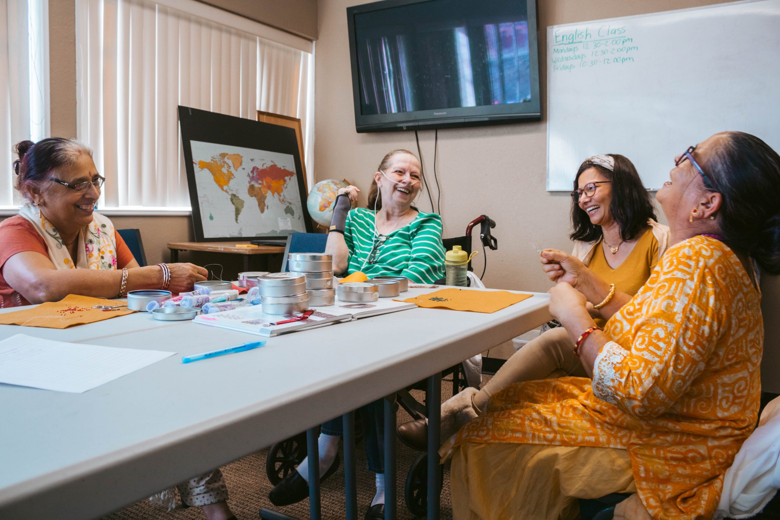 Four individuals are seated around a table in a classroom setting, engaged in a lively discussion with notebooks and cups visible on the table. 