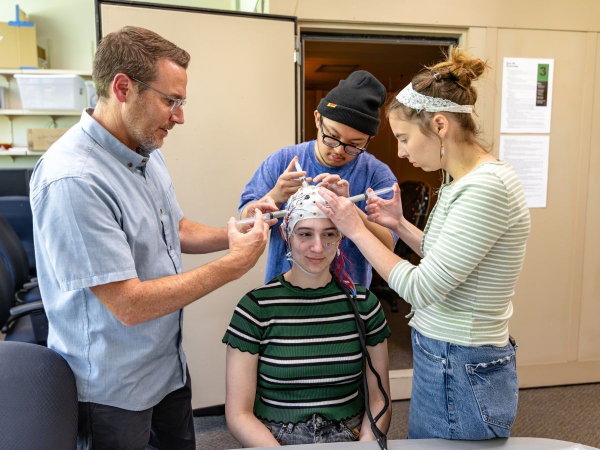 Michael Pitts, a professor at Reed College, and two students use syringes to prepare the electrodes on an EEG cap worn by a seated student in a lab.