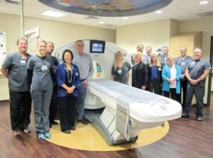 A group of people smile for the camera while gathered around CT-scanner.