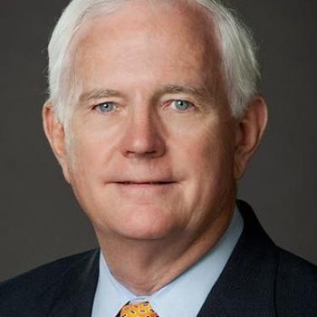 A man with white hair wearing a black suit looks at the camera.