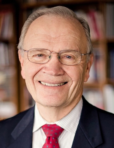 A man with short gray hair wearing glasses and a black suit and red tie smiles for the camera.