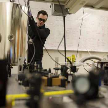 A man with short dark hair wearing dark classes and a black jacket in a lab.