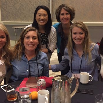 A group of five women gathered around a table smile for the camera.