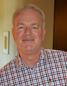 A man with short gray hair wearing a checkered shirt smiles for the camera.