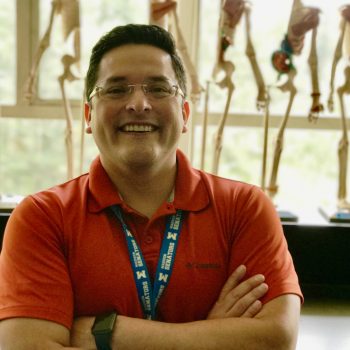 A man with short dark hair wearing a red shirt and glasses with a lanyard around his neck smiles for the camera with his arms crossed.