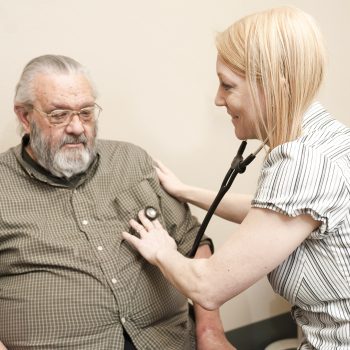 A man with gray hair and facial hair wearing glasses and a checkered shirt gets his heart checked by a woman with straight blond hair wearing a striped hisrt.