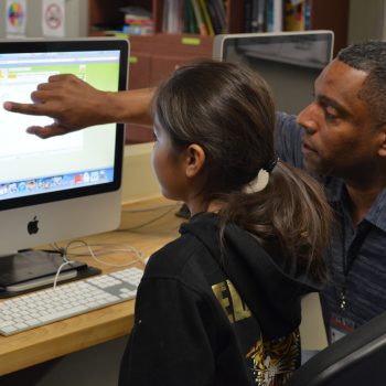 A photo from the Native American Youth and Family Center that shows a man pointing to something on a computer screen next to a young girl with straight dark hair.