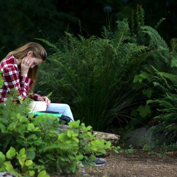 A young woman with straight brown hair wearing a plaid shirt and jeans reads a book while sitting on a bench surrounded by bushes.