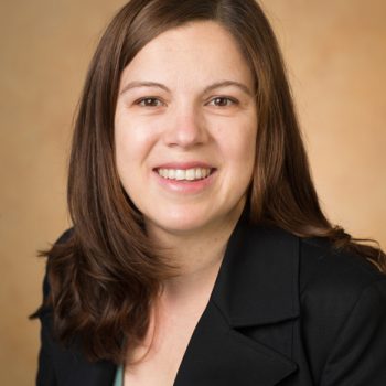 A woman with straight brown hair wearing a black blazer smiles for the camera.