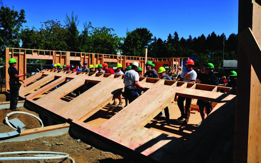 A crew of people wearing hard hats raise a wall on a construction site.