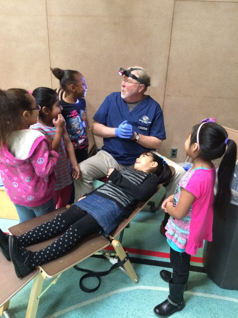 A young girl reclines on a wooden chair while an adult wearing a headlamp explains something to four other young girls.