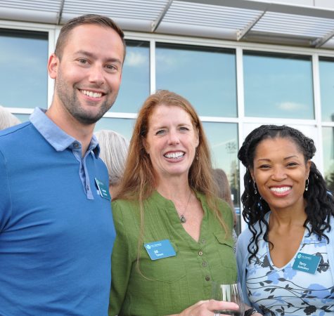 A man with short dark hair wearing a blue shirt, a woman with long red hair wearing a green shirt, and woman with dark hair wearing a blue and white shirt smile for the camera.